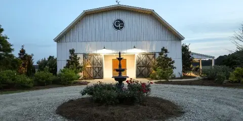 Exterior view of a white barn event venue with a fountain and gravel driveway at dusk.