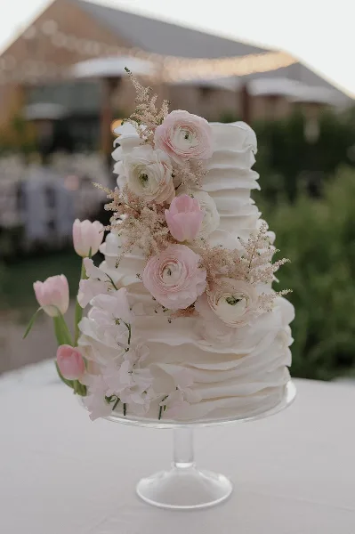 A white wedding cake with ruffled frosting and fresh pink and white floral decorations on a glass stand.