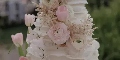 A white wedding cake with ruffled frosting and fresh pink and white floral decorations on a glass stand.
