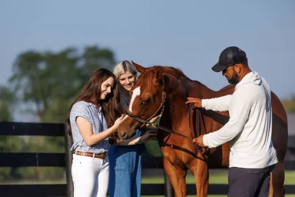 A guide and two guests interacting with a brown horse at a farm.