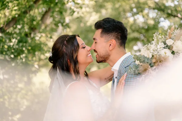 A bride and groom smiling and touching noses in an outdoor setting with green foliage and a floral arrangement.