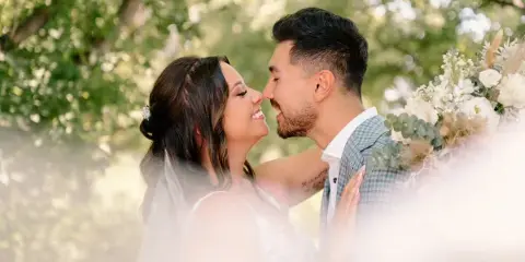 A bride and groom smiling and touching noses in an outdoor setting with green foliage and a floral arrangement.