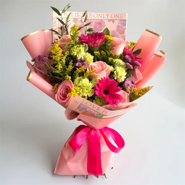 A colorful floral bouquet featuring pink roses, gerbera daisies, and yellow filler flowers, wrapped in pink paper with a pink ribbon.