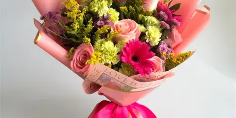 A colorful floral bouquet featuring pink roses, gerbera daisies, and yellow filler flowers, wrapped in pink paper with a pink ribbon.