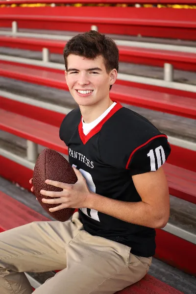 A young man in a black football jersey holding a football, sitting on red stadium bleachers.