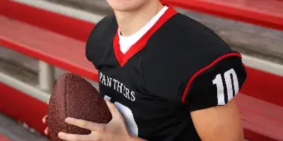 A young man in a black football jersey holding a football, sitting on red stadium bleachers.