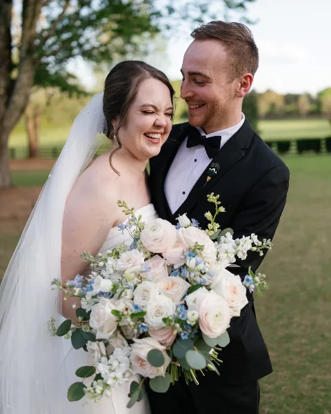 A bride and groom smiling while holding a white and blue wedding bouquet outdoors.
