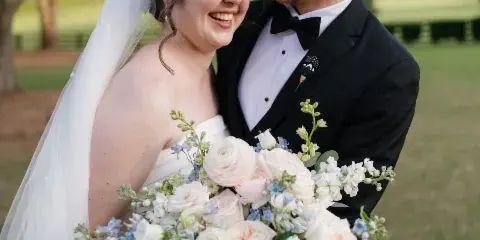 A bride and groom smiling while holding a white and blue wedding bouquet outdoors.