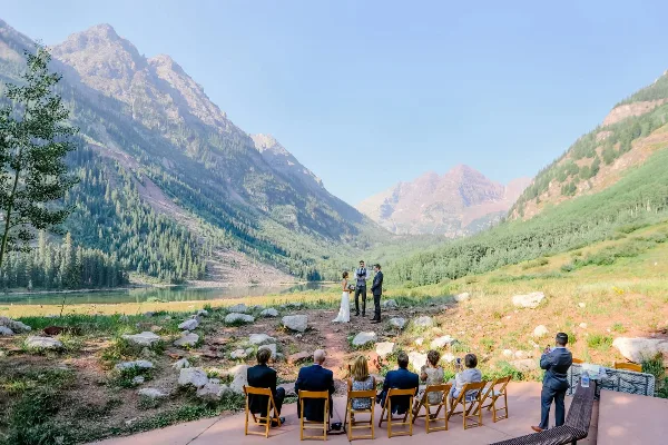 A small wedding ceremony taking place outdoors in a mountain valley with a lake in the background.