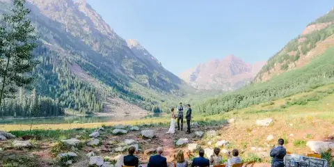 A small wedding ceremony taking place outdoors in a mountain valley with a lake in the background.