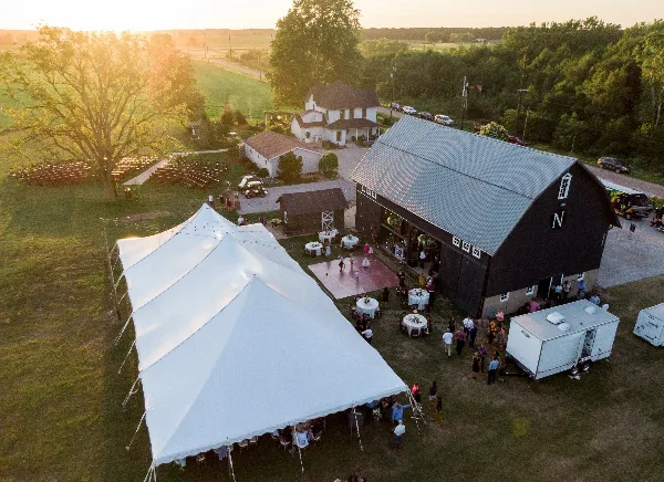 Aerial view of a rustic wedding venue featuring a black barn, white reception tent, and outdoor ceremony seating on a farm property.