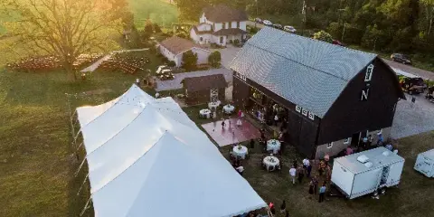 Aerial view of a rustic wedding venue featuring a black barn, white reception tent, and outdoor ceremony seating on a farm property.