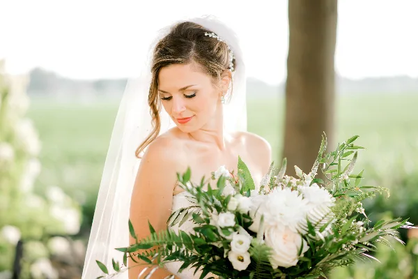 A bride with styled hair and makeup holding a white floral bouquet outdoors.