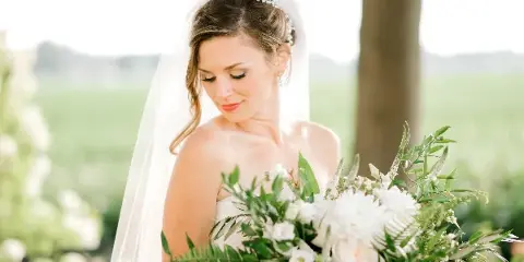 A bride with styled hair and makeup holding a white floral bouquet outdoors.