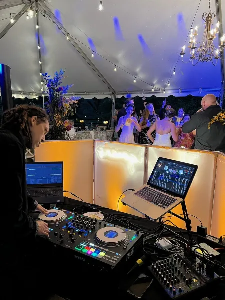 A DJ mixing music on a Serato controller at a wedding reception under a tent with guests dancing in the background.