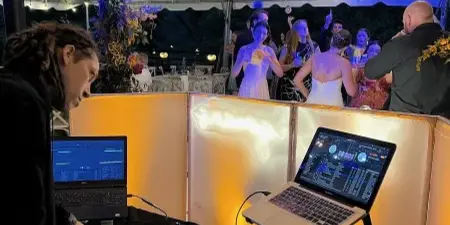 A DJ mixing music on a Serato controller at a wedding reception under a tent with guests dancing in the background.