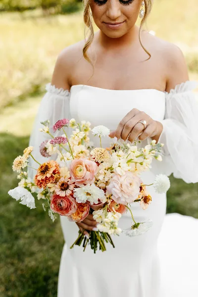 A bride in a white dress holding a colorful wedding bouquet of ranunculus, zinnias, and other fresh flowers.