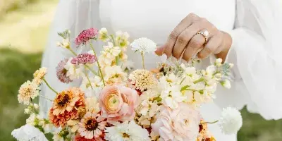 A bride in a white dress holding a colorful wedding bouquet of ranunculus, zinnias, and other fresh flowers.