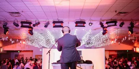 A DJ performing at a crowded event venue with stage lighting and a large audience.