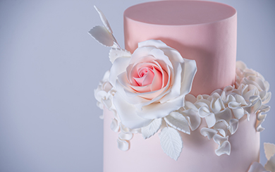 A close-up of a pink two-tier wedding cake decorated with a large white and pink sugar rose and delicate sugar leaves.