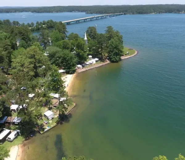 Aerial view of the Children's Harbor Lake Martin campus, showing a wooded peninsula with a white church steeple and buildings situated on a sandy shoreline next to a large lake.