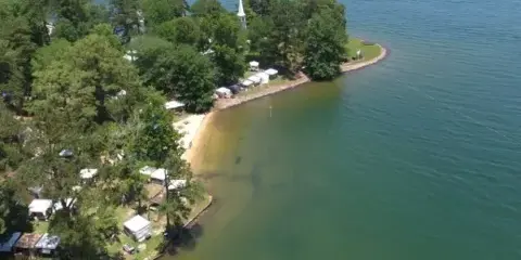 Aerial view of the Children's Harbor Lake Martin campus, showing a wooded peninsula with a white church steeple and buildings situated on a sandy shoreline next to a large lake.