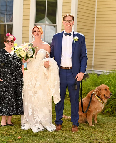 A bride in a white lace dress and a groom in a blue suit stand with a wedding party and a golden retriever in front of a yellow building.