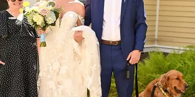 A bride in a white lace dress and a groom in a blue suit stand with a wedding party and a golden retriever in front of a yellow building.
