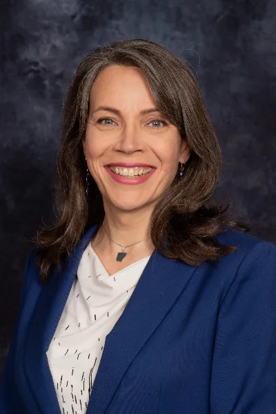 A professional headshot of a smiling woman with shoulder-length brown hair, wearing a blue blazer and a white patterned top.