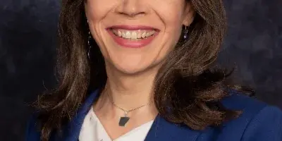 A professional headshot of a smiling woman with shoulder-length brown hair, wearing a blue blazer and a white patterned top.