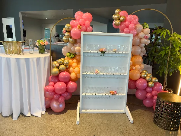 A decorated event space featuring a white table with linens, a glassware rack, and pink and gold balloon arches.