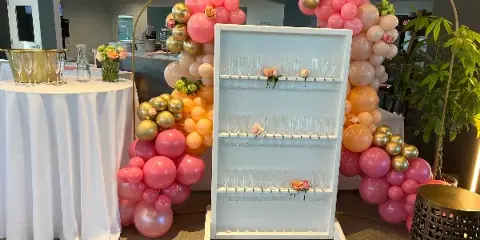 A decorated event space featuring a white table with linens, a glassware rack, and pink and gold balloon arches.