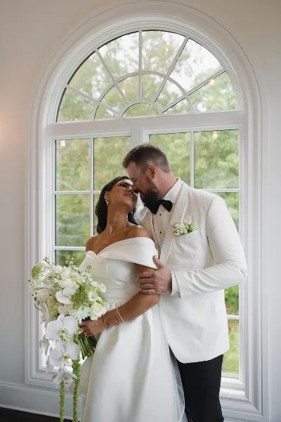 A bride in a white gown holding a cascading bouquet of white orchids and roses while standing with a groom in a white tuxedo.