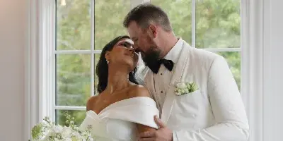 A bride in a white gown holding a cascading bouquet of white orchids and roses while standing with a groom in a white tuxedo.