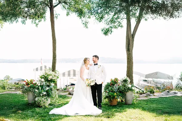 A bride and groom standing together outdoors near a lake, with the bride holding a bouquet and the groom in a white tuxedo jacket.