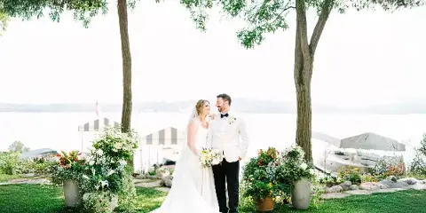 A bride and groom standing together outdoors near a lake, with the bride holding a bouquet and the groom in a white tuxedo jacket.