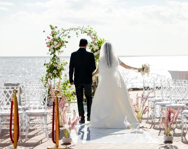 A bride and groom walking down an aisle toward a floral arch at a seaside wedding venue.