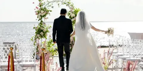 A bride and groom walking down an aisle toward a floral arch at a seaside wedding venue.