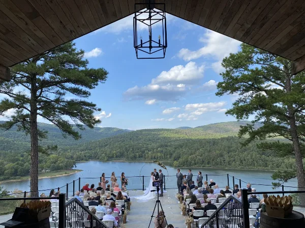 A wedding ceremony in progress on an outdoor deck overlooking a lake and forested hills.