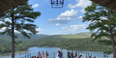 A wedding ceremony in progress on an outdoor deck overlooking a lake and forested hills.