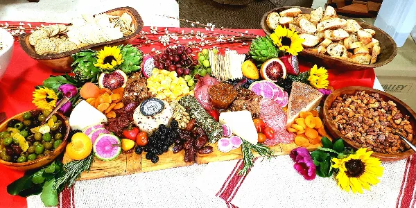 A large, colorful catering spread on a wooden board featuring an assortment of cheeses, cured meats, fresh fruits, nuts, and breads, garnished with sunflowers.