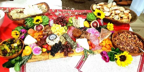 A large, colorful catering spread on a wooden board featuring an assortment of cheeses, cured meats, fresh fruits, nuts, and breads, garnished with sunflowers.