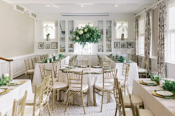 An elegant event venue interior featuring round tables with white linens, gold chiavari chairs, and a large floral centerpiece.