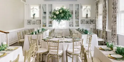 An elegant event venue interior featuring round tables with white linens, gold chiavari chairs, and a large floral centerpiece.