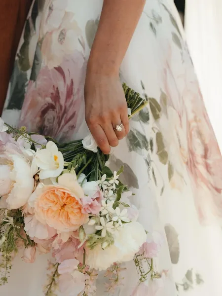 A bride in a floral dress holding a bouquet of white and peach roses and greenery.