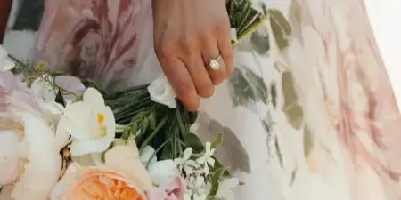 A bride in a floral dress holding a bouquet of white and peach roses and greenery.