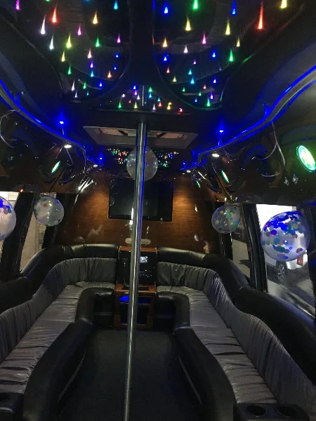 Interior of a party bus featuring a central dance pole, colorful ceiling lights, and black leather bench seating.