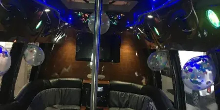 Interior of a party bus featuring a central dance pole, colorful ceiling lights, and black leather bench seating.