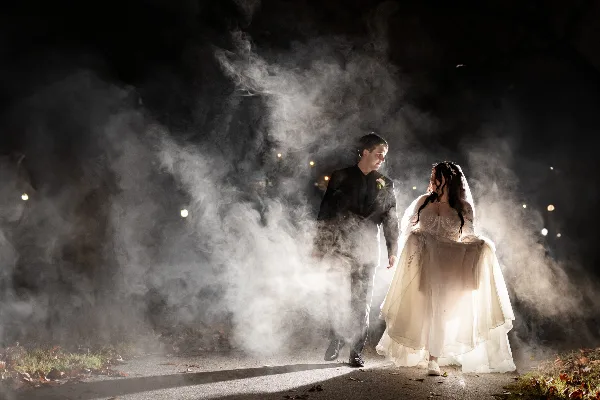 A bride and groom walking through a cloud of white smoke at night.