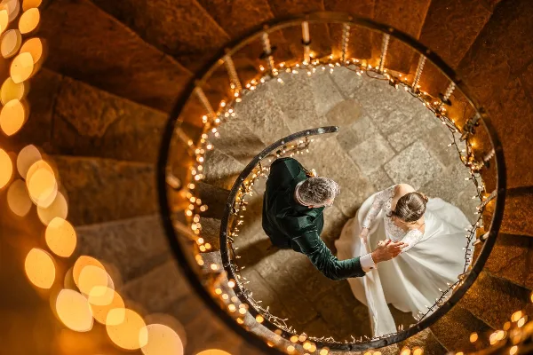 A bride and groom holding hands on a spiral staircase decorated with string lights, viewed from above.